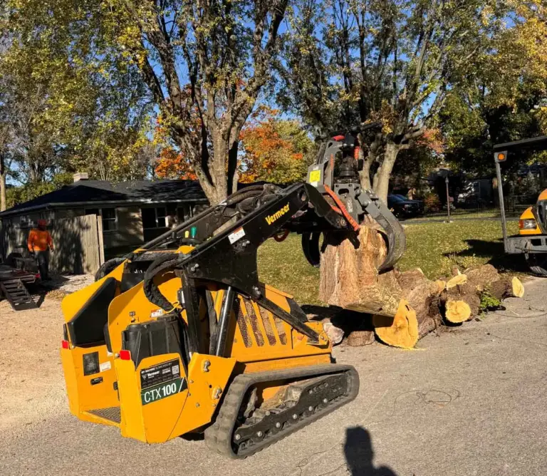 Tracked loader with a grapple picking up a log on a driveway